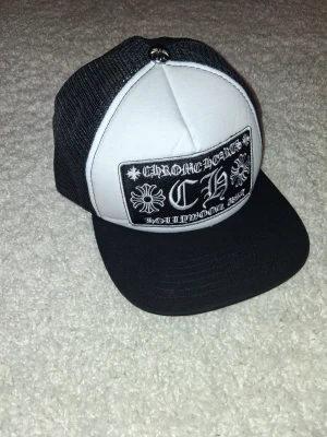 Svartvit keps från Chrome Hearts - Säljer en svartvit truckerkeps från Chrome Hearts med stort broderat märke framtill där det står 'CH' och 'Hollywood, USA'. Kepsen har svart skärm, vit front och svart mesh baktill för extra ventilation. Perfekt för dig som gillar streetwear och vill sticka ut.