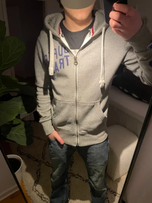 Grå zip-hoodie från Superdry - Snygg grå hoodie med dragkedja från Superdry. Har blå tryck på bröstet och längs ena ärmen, samt vita dragsnören i huvan. Mjuk insida och två fickor framtill. Perfekt för en avslappnad streetstyle-look.
