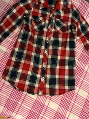 Rutig skjorta från Polarn O. Pyret - Röd, blå och vit rutig skjorta från Polarn O. Pyret med långa ärmar och knappar framtill. Skjortan har två bröstfickor med lock och klassisk krage med blommigt mönster på insidan. Perfekt för en avslappnad och cool stil.