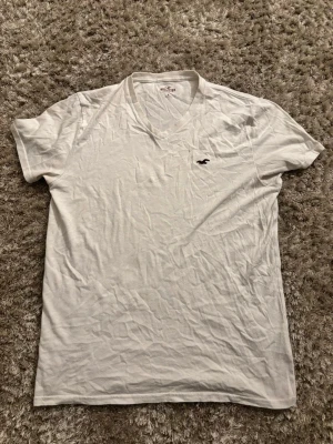 Vit v-ringad t-shirt från Hollister - Klassisk vit t-shirt från Hollister med v-ringning och korta ärmar i storlek M.Tillverkad i mjuk bomull och har Hollisters ikoniska logga broderad på bröstet. Enkel och stilren design som passar till det mesta. Har du några funderingar så skriv gärna👍