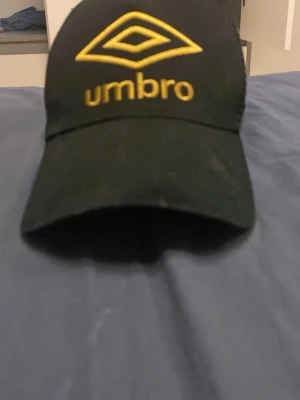 Svart keps från Umbro med gul logga - Snygg svart keps från Umbro med stor gul broderad logga och text framtill. Klassisk böjd skärm och mesh baktill för extra ventilation. Justerbar passform med snapback-spänne. Perfekt för en sportig och avslappnad stil. Ser fläckig ut men är bättre i verkligheten 