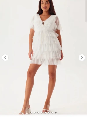 Studentklänning  - Superfin kort vit klänning med flera lager volanger och transparenta korta ärmar. Djup v-ringning med knyt fram och öppen rygg med knytband. Använd endast på studenten så är i väldigt bra skick! 