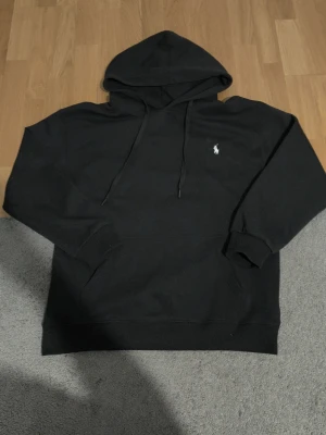 Svart hoodie från Polo Ralph Lauren - Svart hoodie från Polo Ralph Lauren med klassisk huva och känguruficka. Aldrig använd, nypris 1200. Storlek är XL men passar L. Ikoniska vita Polo-loggan broderad på bröstet. Mjuk bomullsblandning och ribbade muddar vid ärmslut och nederkant. Perfekt för en avslappnad och stilren look.