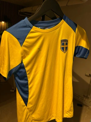 Sverige matchtröja gul/blå - Gul och blå matchtröja från Adidas med Sveriges landslagsemblem på bröstet. Tröjan har korta ärmar, rund hals och är tillverkad i ett lätt och ventilerande material som passar perfekt till träning eller match.