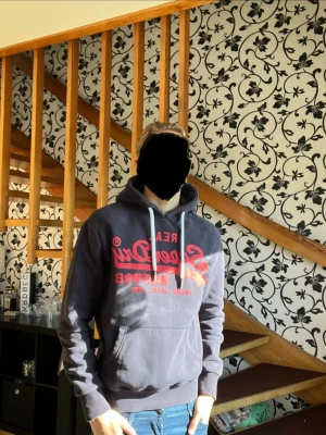Mörkblå Superdry hoodie med tryck - Mörkblå hoodie från Superdry med stort rött tryck på bröstet och vita dragsnören i huvan. Klassisk känguruficka framtill och ribbade muddar vid ärmslut och midja. Perfekt för en avslappnad streetstyle-look.