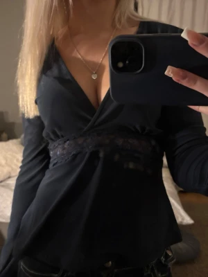 Svart omlottopp med spetsdetalj - Svart omlottopp med v-ringning och spetsdetalj under bysten. Långärmad modell som sitter snyggt och markerar midjan. Perfekt att styla med jeans eller kjol.