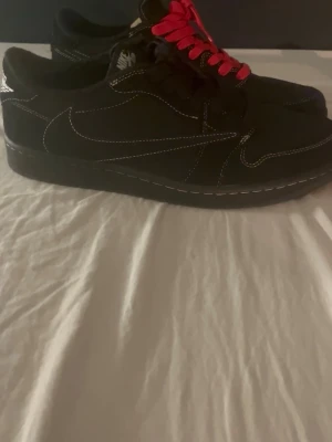 Nike Air Force 1 Low x Travis Scott “Black” - Svarta Nike Air Force 1 Low sneakers med kontrasterande vita sömmar och röda skosnören. Klassisk siluett med Nike-logga på sidan och bak. Ovandel i mocka för en stilren och trendig look.