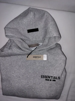 Grå Essentials Fear of God hoodie - Snygg grå hoodie från Essentials Fear of God med svart logga tryckt på bröstet. Klassisk passform med huva och ribbade muddar. Tillverkad i mjuk bomullsblandning som känns skön mot huden. Perfekt för en avslappnad och trendig streetwear-look.
