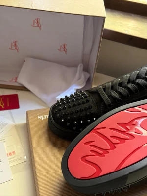 Christian Louboutin svarta sneakers med nitar - Svarta sneakers från Christian Louboutin med mattsvarta nitar på tån och klassisk röd sula med logotyp. Skorna har svarta skosnören och är tillverkade i mocka och läder. Snygg och edgy design som sticker ut.
