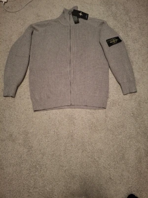 Grå ziptröja från Stone Island - Säljer en grå ribbstickad ziptröja från Stone Island med hög krage och ikonisk patch på ärmen. Tröjan har hel dragkedja framtill och ribbade muddar vid ärmslut och nederkant. Perfekt för dig som gillar stilrena och exklusiva plagg.