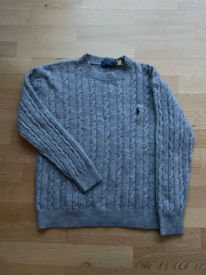Grå kabelstickad tröja Polo Ralph Lauren - Kabelstickad grå tröja från Polo Ralph Lauren med rund halsringning och broderad logga på bröstet. Tillverkad i mjuk ullblandning och har ribbade muddar vid ärmslut och nederkant. Perfekt för lager på lager och klassisk stil.