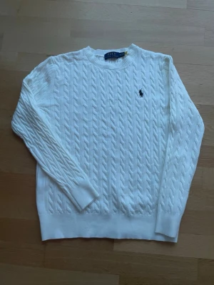 Vit kabelstickad tröja Polo Ralph Lauren - Kabelstickad vit tröja från Polo Ralph Lauren med rund halsringning och den klassiska broderade loggan på bröstet. Tröjan har långa ärmar och ribbade muddar vid ärmslut och nederkant. Perfekt för en clean och stilren look.