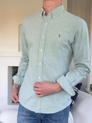 Grön skjorta från Polo Ralph Lauren (Slim Fit) - Klassisk ljusgrön skjorta från Polo Ralph Lauren med button-down krage och broderad logga på bröstet. Skjortan har långa ärmar, knäppning framtill och är tillverkad i bomull. Perfekt för en stilren och avslappnad look. Storlek Medium kan passa small. Snabb leverans!