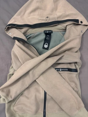 Beige hoodie från Sail Racing - Snygg beige hoodie från Sail Racing med dragkedja framtill och svart logotypdetalj på bröstet. Hoodien har en stor huva med tryckknappar och meshfoder på insidan för extra komfort. Perfekt för en avslappnad och sportig stil. Storlek XS