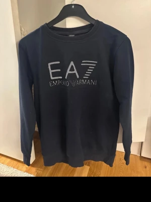 Svart sweatshirt från Emporio Armani - Svart sweatshirt från Emporio Armani, modell EA7, med broderad logga i vitt på bröstet. Tröjan har rund hals och långa ärmar, tillverkad i mjukt material som känns skönt mot huden. Perfekt för dig som gillar stilrena och exklusiva märken.