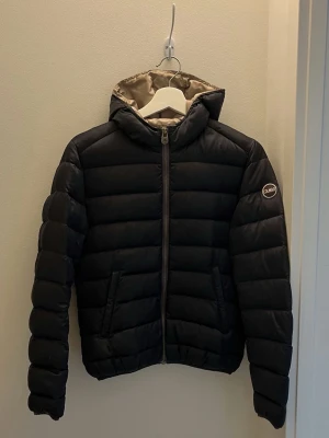 Mörkblå dunjacka från Colmar - Snygg mörkblå dunjacka från Colmar Originals med quiltad design och huva. Jackan har dragkedja framtill, två fickor med dragkedja och Colmar-logga på ärmen. Insidan är ljusgrå och materialet är lätt men varmt, perfekt för kalla dagar. Inga defekter för dam. Passar liten S/XS