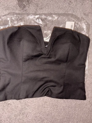 Svart axelbandslös tubtopp - Snygg svart axelbandslös tubtopp med en v-formad urringning framtill. Toppen är croppad och har en stilren, tight passform. I originalförpackning samt med prislapp. Säljer då jag missa returtiden. Ursprungspris 399 kr från NA-KD.