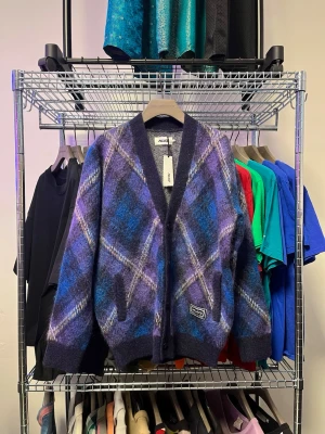 Lila-rutig kofta från Palace - Säljer en fluffig kofta från Palace med rutigt mönster i lila, blått och svart. Koftan har v-ringad öppning, ribbade muddar och en patch med logga nedtill. Perfekt för dig som gillar streetwear och vill sticka ut med färg och mönster.