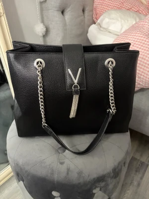 Valentino väska - Snygg svart handväska i konstläder med silverfärgade kedjehandtag och dekorativ V-detalj framtill. Väskan har ett rymligt huvudfack, innerficka med dragkedja och magnetlås.