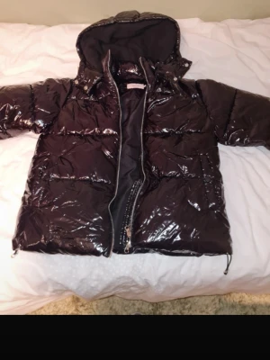  pufferjacka från Nelly - Säljer en marinblå glansig pufferjacka från Nelly med huva och dragkedja framtill. Jackan har en quiltad design och är perfekt för kalla vinterdagar. Materialet är syntetiskt med en blank finish som ger en trendig look.