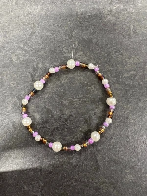 Pärlarmband i olika färger Storlek M  - Ett handgjort armband med vita pärlor, små lila och guldiga glaspärlor på elastisk tråd. Perfekt accessoar för att addera lite färg och stil till din outfit.