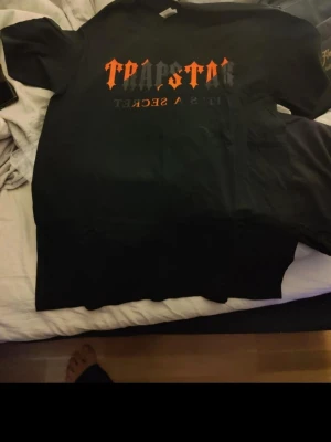 T svart t-shirt med tryck - Svart t-shirt från T med stort tryck i orange och grått på bröstet där det står 'Trapstar' och 'It's a secret'. Klassisk passform och rund hals. Perfekt för dig som gillar streetwear och vill sticka ut med en snygg grafisk tee. Är Shorts och t-shirt finns olika storlekar 