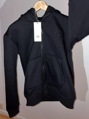 Svart hoodie från C.P. Company - Svart hoodie med dragkedja från C.P. Company. Klassisk design med två fickor framtill och broderad logga på bröstet. Huvan har inbyggda goggles, vilket ger en unik och streetinspirerad look. Perfekt för dig som gillar stilrena och coola plagg.