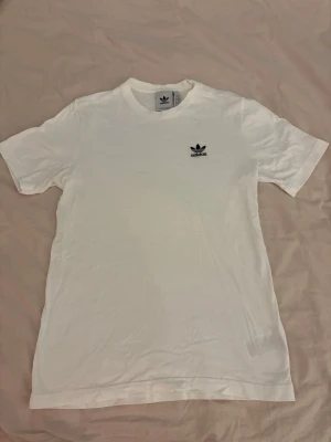 Vit Adidas t-shirt med logga - Klassisk vit t-shirt från Adidas med svart broderad logga på bröstet. T-shirten har rund halsringning och korta ärmar. Perfekt basplagg med stilren design och skön passform.
