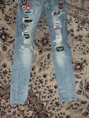 Ljusblå patchade skinny jeans från amiri - Unika ljusblå skinny jeans med slitningar, färgstänk och flera coola patches och broderade märken på framsidan. Jeansen har en trendig distressed look och smal passform. Perfekta för dig som vill sticka ut med din stil.