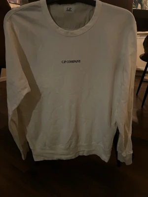 Vit sweatshirt från C.P. Company - Säljer en stilren vit sweatshirt från C.P. Company med rund halsringning och diskret logga på bröstet. Tröjan har långa ärmar och är gjord i mjukt bomullsmaterial. Perfekt för en clean och avslappnad look.