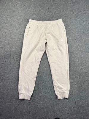 Beige mjukisbyxor från Adidas - Säljer ett par beige mjukisbyxor från Adidas med resår i midjan och muddar vid bensluten. Byxorna har två sidofickor och den klassiska Adidas-loggan på vänster ben. Perfekta för chill dagar eller träning.
