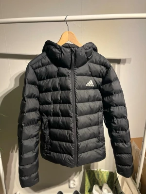 Svart dunjacka från Adidas - Snygg svart dunjacka från Adidas i storlek S med huva och dragkedja framtill. Jackan har quiltad design, två fickor med dragkedja och Adidas-logga på bröstet. Perfekt för kalla vinterdagar och riktigt skön att bära.  Små defekter såsom den sista bilden visar förekommer.