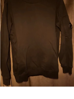 Svart sweatshirt från C.P. Company - Svart sweatshirt från C.P. Company med rund halsringning och lång ärm. Klassisk patch med logga på ena ärmen och ribbade muddar vid ärmslut och nederkant. Perfekt för en clean och stilren look.