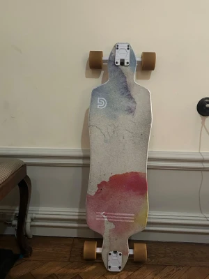 Färgglad longboard från DB Longboards - Säljer en longboard från DB Longboards med unikt färgstarkt mönster i rött, gult och lila på vit botten. Brädan har drop-through trucks och bruna hjul. Perfekt för cruising och carving i stan eller parken. Har repor se bild.