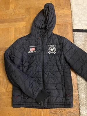 Svart dunjacka Vinson Polo Club - Svart dunjacka från Vinson Polo Club med huva och dragkedja. Jackan har broderade loggor på bröstet och quiltat mönster. Perfekt för kalla vinterdagar och har ribbade muddar vid ärmsluten.