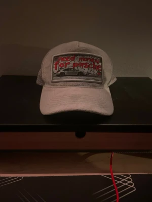 Grå keps med Porsche-tryck - Snygg grå keps med mesh på sidorna och ett stort tygmärke framtill med texten 'need money for porsche' och en bild på en bil. Kepsen har böjd skärm och justerbar baksida. Perfekt accessoar för dig som gillar bilar och streetwear.