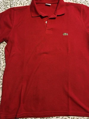  Lacoste Polo Shirt (RARE) - Klassisk röd pikétröja från Lacoste med broderad logga på bröstet. Tröjan har krage, korta ärmar och knappar vid halsen. Perfekt för en stilren och sportig look.
