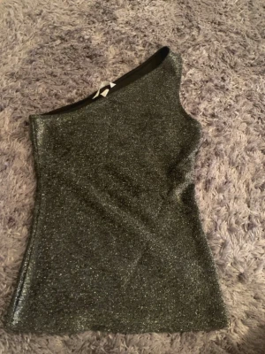 Glittrig one shoulder topp - Snygg glittrig topp i silver med one shoulder-design. Toppen är figurnära och har en axelbandslös look på ena sidan, vilket ger en trendig och unik stil. Perfekt för dig som vill sticka ut med lite extra glitter.