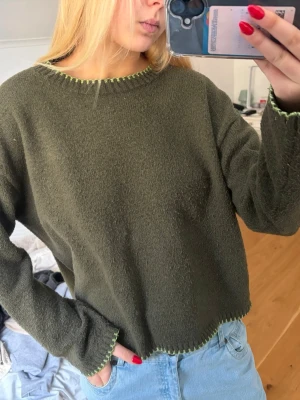 Olivgrön stickad tröja med kontrastsöm - Mysig olivgrön stickad tröja med rund hals och dekorativ ljusgrön kontrastsöm vid hals, ärmslut och nederkant. Tröjan har en avslappnad passform och långa ärmar. Perfekt att matcha med jeans för en chill look.