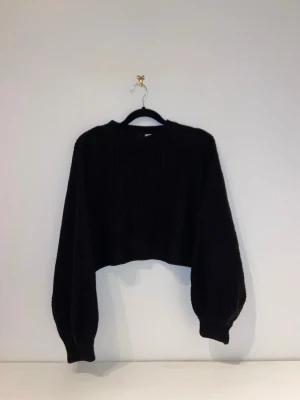 Svart stickad tröja - Säljer en svart stickad tröja med ballongärmar och ribbade muddar. Tröjan har en enkel, stilren design och är perfekt för lager på lager. Passar dig som gillar trendiga och bekväma plagg.