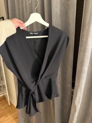 Svart omlottopp från Zara - Snygg svart omlottopp från Zara med knyt i midjan och v-ringad hals. Toppen är ärmlös och har en figurnära passform som framhäver siluetten. Perfekt att styla med jeans eller kjol för en trendig look.