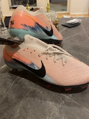 Nike united mercurial vapor 16 elite - Snygga Nike Mercurial fotbollsskor i rosa och beige med svarta detaljer och blå inslag. Skorna har en strömlinjeformad design, stickad ovandel och klassisk snörning. Perfekta för dig som vill sticka ut på planen och få grym känsla med bollen.