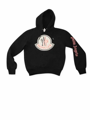 Svart Moncler x Palm Angels hoodie - Svart hoodie med stor Moncler-logga framtill och Palm Angels-tryck i rosa på ena ärmen. Klassisk huva med dragsko och ribbade muddar. Snygg streetwear-stil, perfekt för dig som gillar exklusiva samarbeten och tydliga loggor.