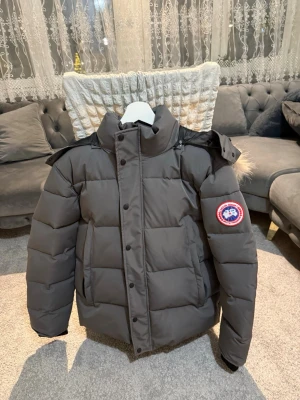  dunjacka från Canada Goose M/L-XL - Säljer en Grå dunjacka från Canada Goose med klassisk patch på ärmen. Jackan har hög krage, avtagbar huva med pälskant och stora framfickor. Perfekt för kalla vinterdagar och riktigt varm tack vare dunfyllningen. Stängs med dragkedja och knappar. PASSAR 