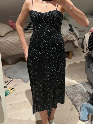 Svart prickig midiklänning med smala band - Säljer svart klänning med vita prickar och smala axelband. Klänningen har en figurnära passform, öppen rygg med knyt och är tillverkad i ett glansigt material som ger en lyxig känsla. 