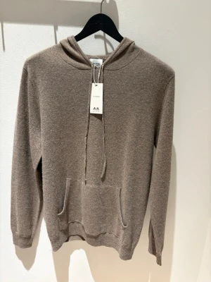 Beige cashmere hoodie John Henric - Mjuk och lyxig beige hoodie från John Henric i 100% cashmere. Klassisk modell med huva, dragsko och känguruficka framtill. Perfekt för dig som vill ha en stilren och bekväm look med exklusiv känsla.