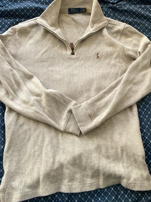 Beige halvzip från Polo Ralph Lauren - Stilren beige tröja med halv dragkedja och hög krage från Polo Ralph Lauren. Tröjan har lång ärm, diskret ribbad struktur och den klassiska broderade loggan på bröstet. Perfekt för en avslappnad men snygg look.