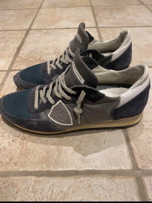 Philippe Model sneakers i blått och grått - Snygga Philippe Model sneakers i en mix av blått, grått och vitt. Skorna har rund tå, snörning och detaljer i mocka och skinn. Klassisk design med logotyp på sidan och bekväm platt sula. Perfekta för dig som gillar stilrena sneakers med premiumkänsla.