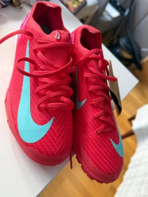 Nike fotbollsskor i rött och turkos - Snygga fotbollsskor från Nike i en stark röd färg med turkosa swoosh-detaljer. Skorna har snörning och är tillverkade i ett syntetmaterial som ger bra grepp och känsla på planen. Perfekta för dig som vill sticka ut på fotbollsplanen.