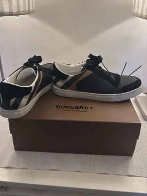 Burberry låga sneakers i svart och beige - Snygga låga sneakers från Burberry i svart textil och läder med klassiskt rutmönster i beige, vitt och svart på sidorna. Skorna har vita sulor och svarta skosnören. Perfekta för dig som vill ha en stilren och lyxig look. Dustbag och burberry kort till kommer samt låda 